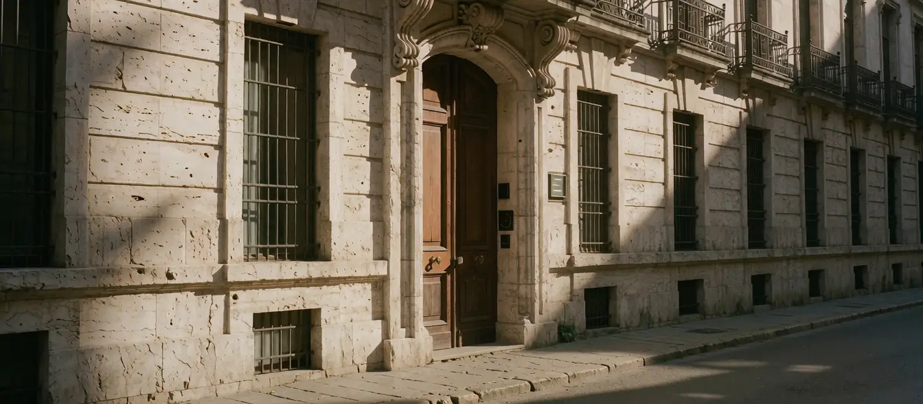 Fachada de piedra de un edificio consular con puertas de madera y balcones de hierro, a la luz de la tarde.